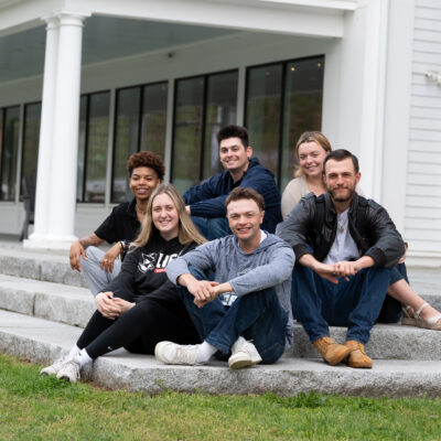 A diverse group of New England College students sit on the steps of the John Lyons Building on campus.