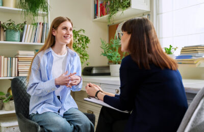A counselor listens to a teenage client in an office lined with books and plants.